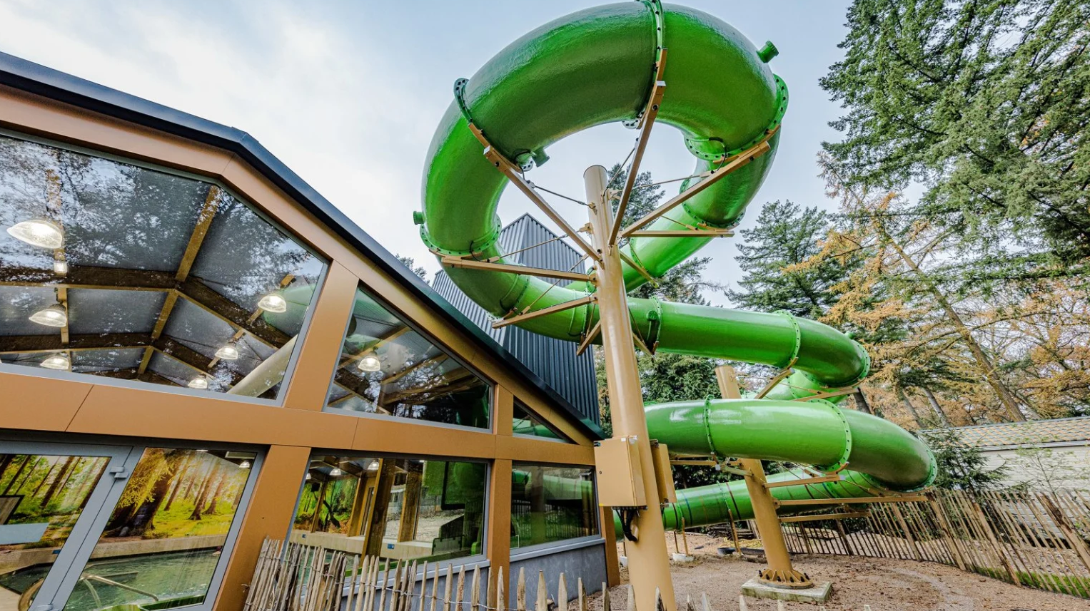 A green water slide spirals outside a wooden building amid tall trees with clear skylights at Europarcs De Utrechtse Heuvelrug