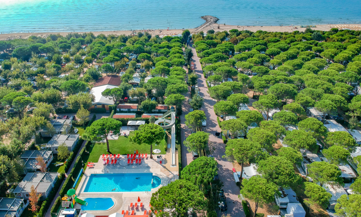 Aerial view of a campsite with swimming pool, scattered trees, and nearby beach, surrounded by lush greenery at Italy Camping Village