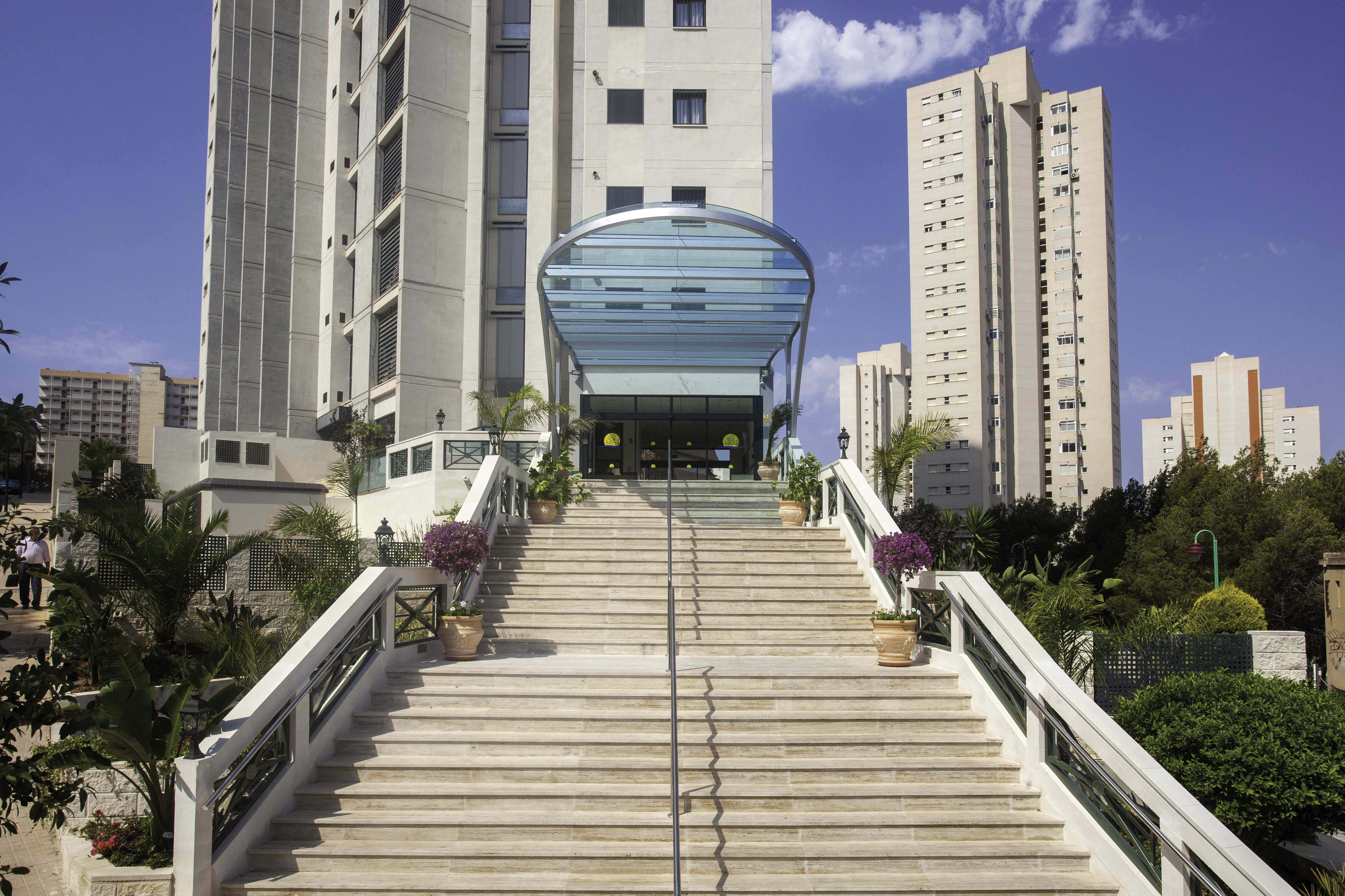 A wide staircase with plants ascends to a building entrance with a glass canopy; tall buildings surround in an urban area at Benidorm Horizon