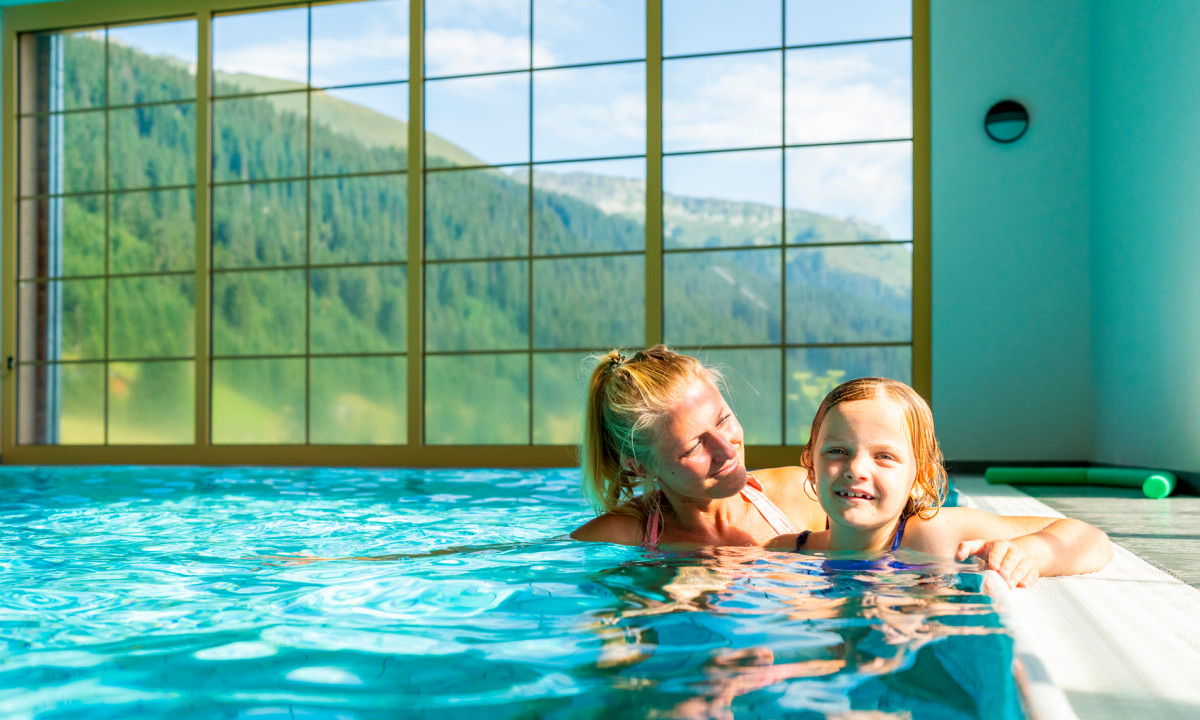Two people swimming in an indoor pool with large windows showcasing mountain scenery at Landal Hochmontafon