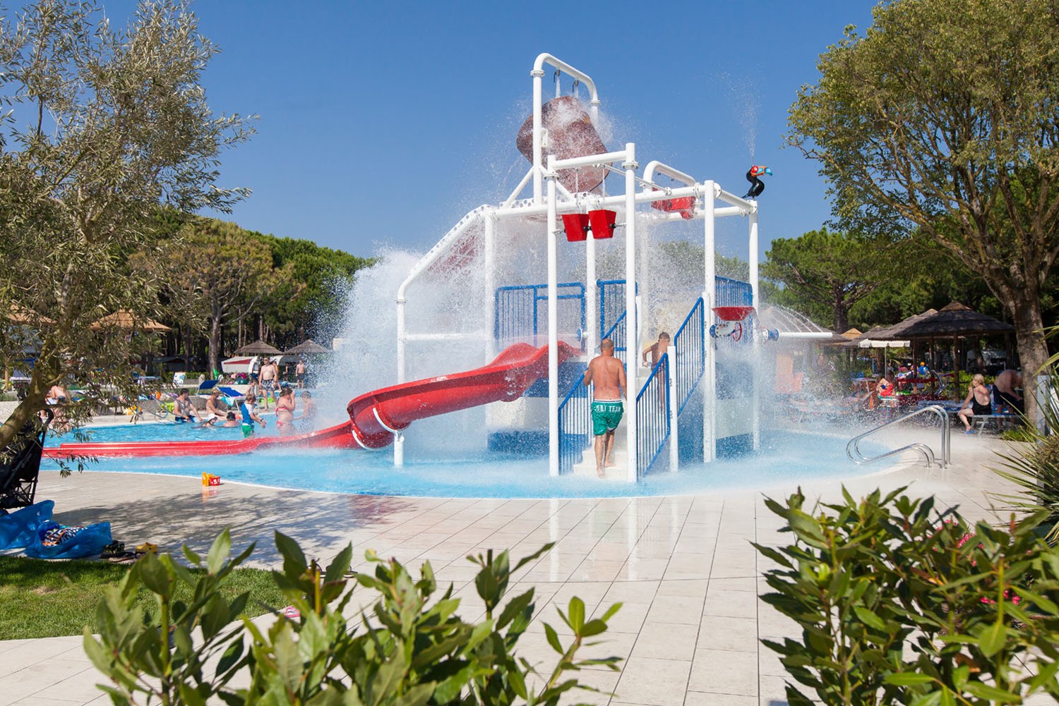 Water play structure releasing splash, children and adults enjoying in pool area, surrounded by trees and greenery at Ca'Pasquali