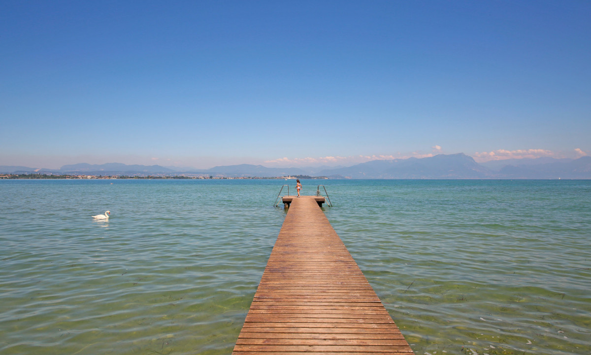 A wooden dock extends into the clear lake, where a swan swims, with mountains and blue sky in the background at Tiglio