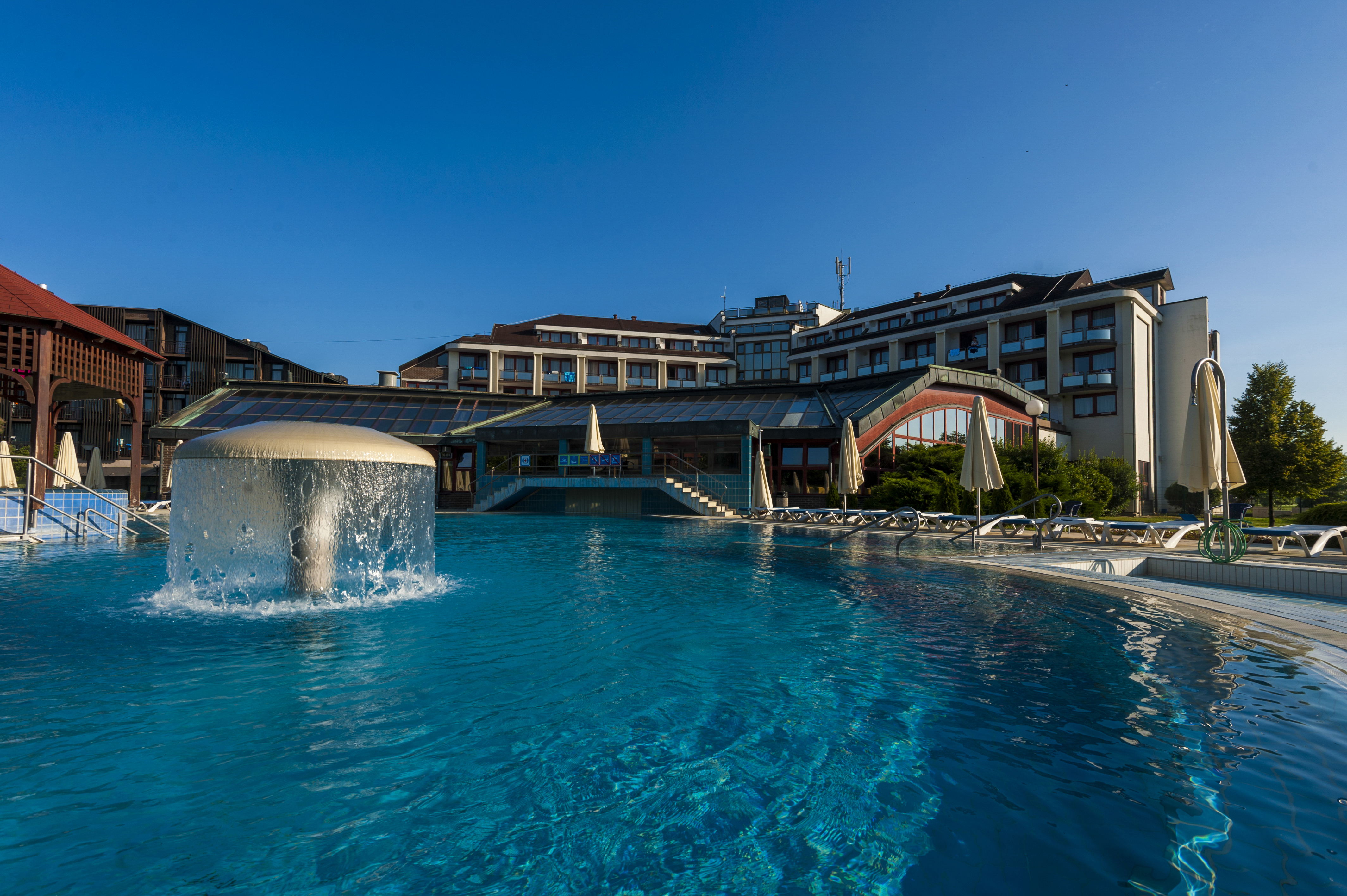 A large pool features a central, umbrella-shaped fountain; a hotel building is visible in the background at Hotel Ajda