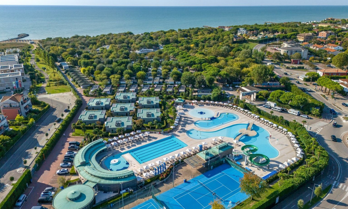 Swimming pools surrounded by lounge chairs, adjacent tennis courts, trees, buildings, and coastline visible in the distance at Europa Camping Village