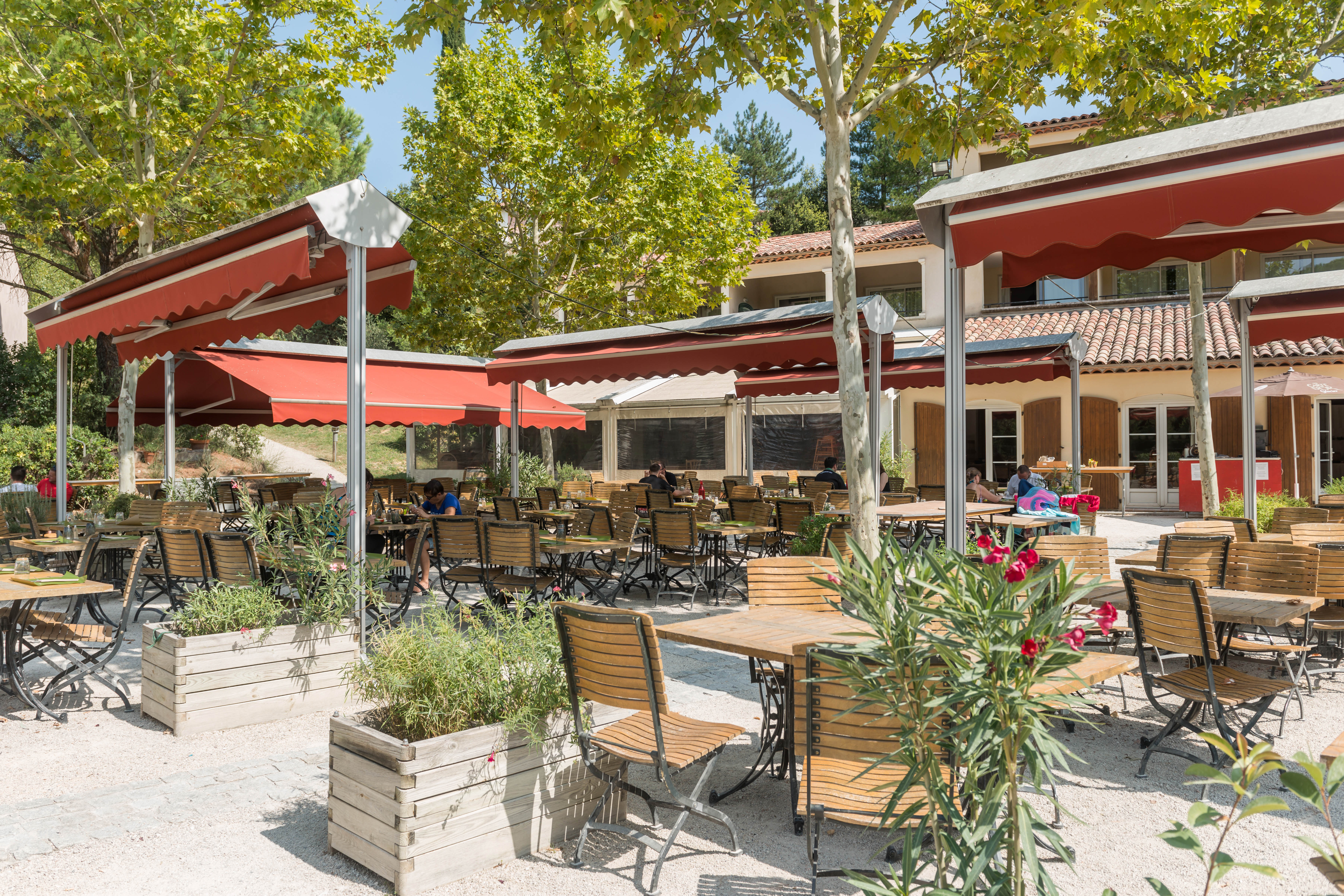 Wooden tables and chairs under red awnings sit empty outside a building with trees and people around at Le Rouret en Ardeche