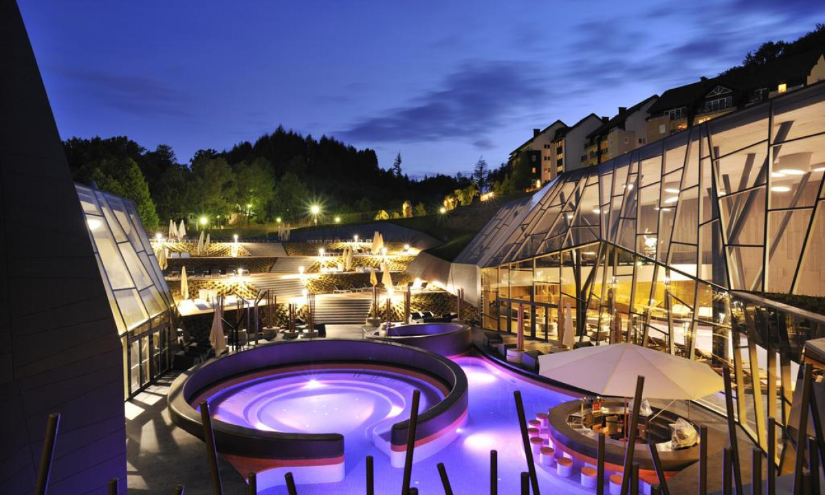Lit pool with circular design, surrounded by modern buildings and bright outdoor lighting, against a twilight forest background at Hotel Sotelia