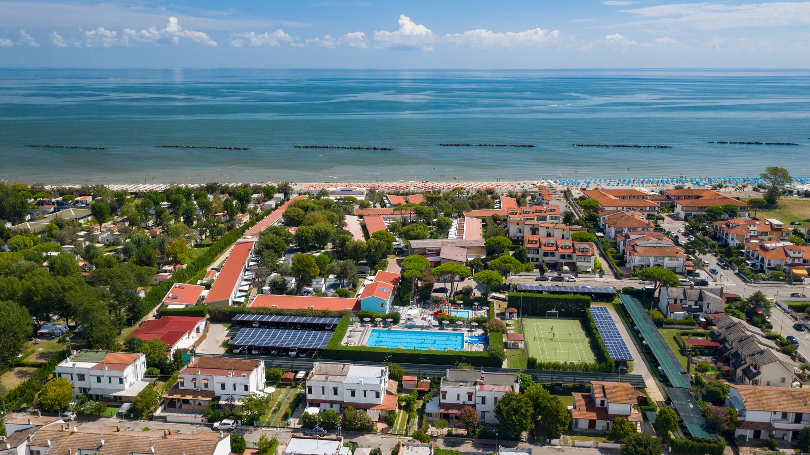 Beachfront campsite surrounded by lush greenery and orange-roofed buildings, with swimming pools, tennis courts, and nearby residential area at Park Gallanti Holiday Village