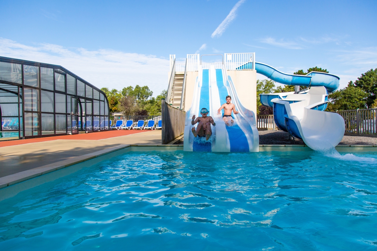 Two individuals splash into a swimming pool from dual water slides beside an enclosed pool area at Domaine de Bréhadour