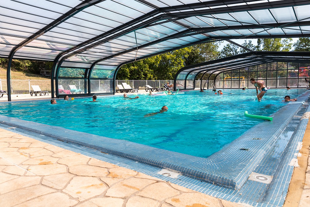 People swimming and lounging at a covered pool area surrounded by sun loungers and trees at Le Saint Louis