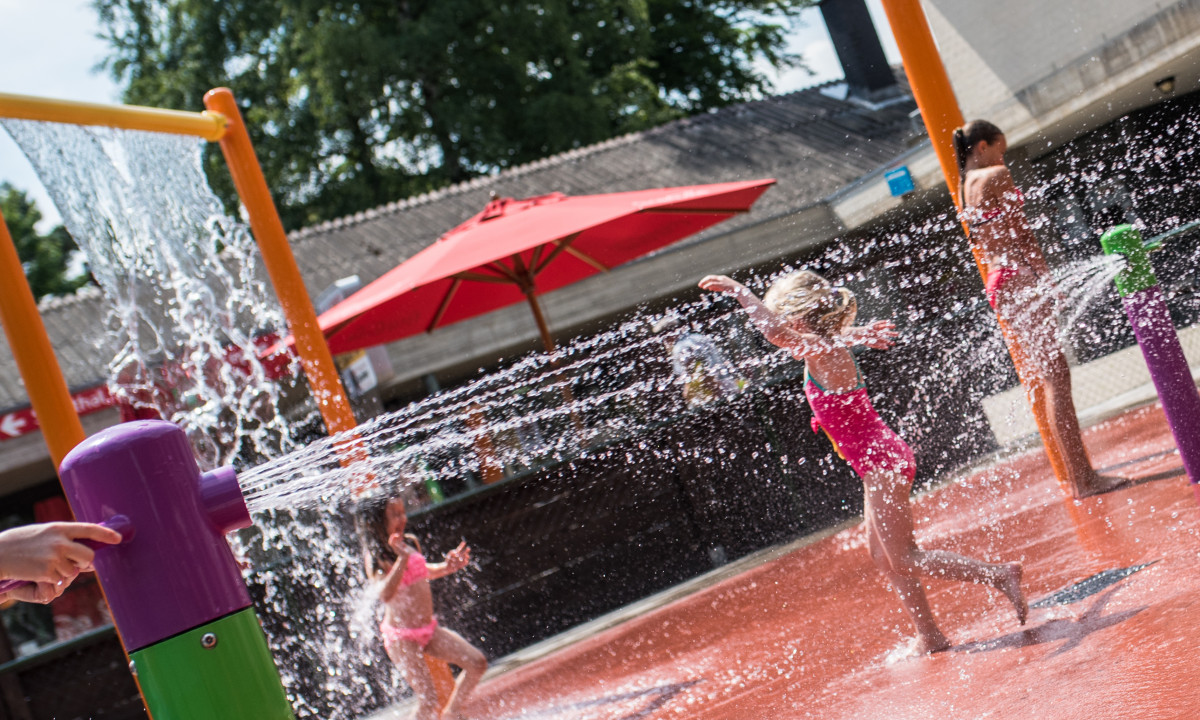 Children playing in a water park with colorful sprinklers and red umbrellas at het Veen