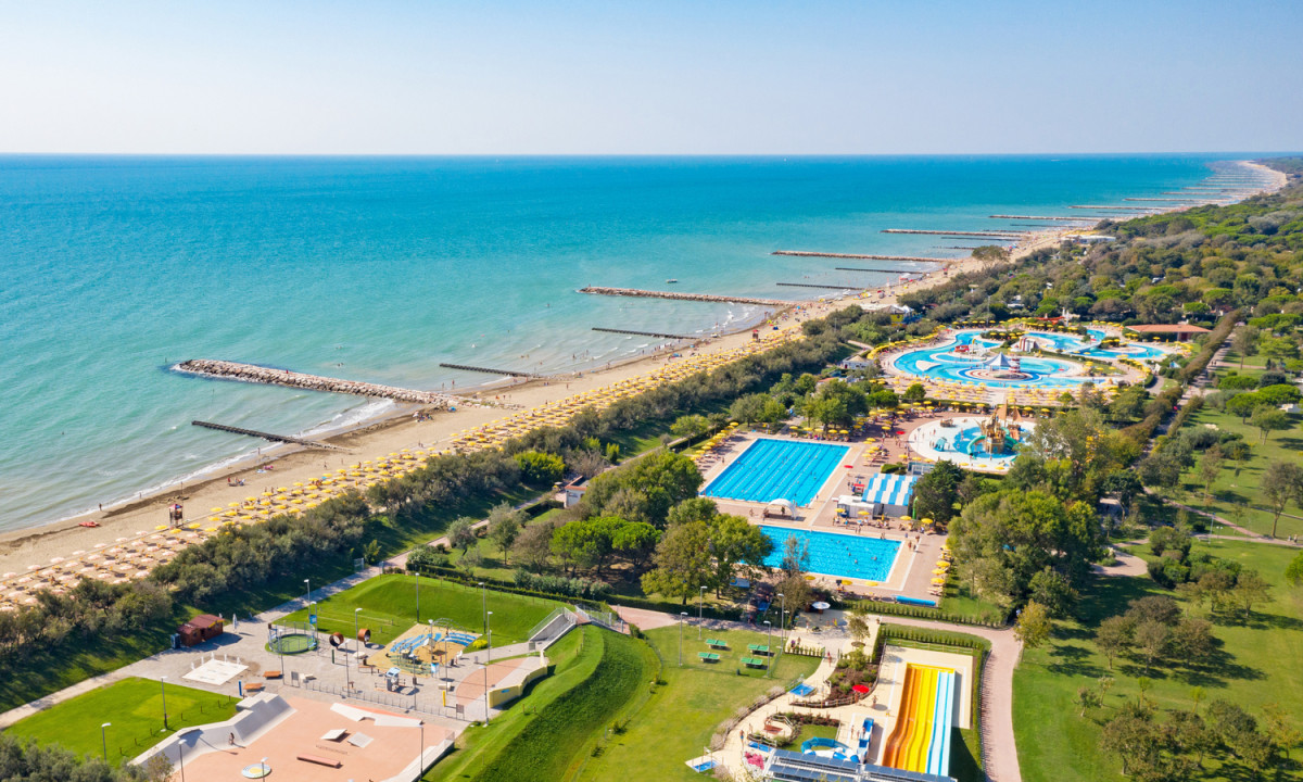 Aerial view of beachgoers under yellow umbrellas, adjacent to a sprawling water park and green recreational area at Pra delle Torri