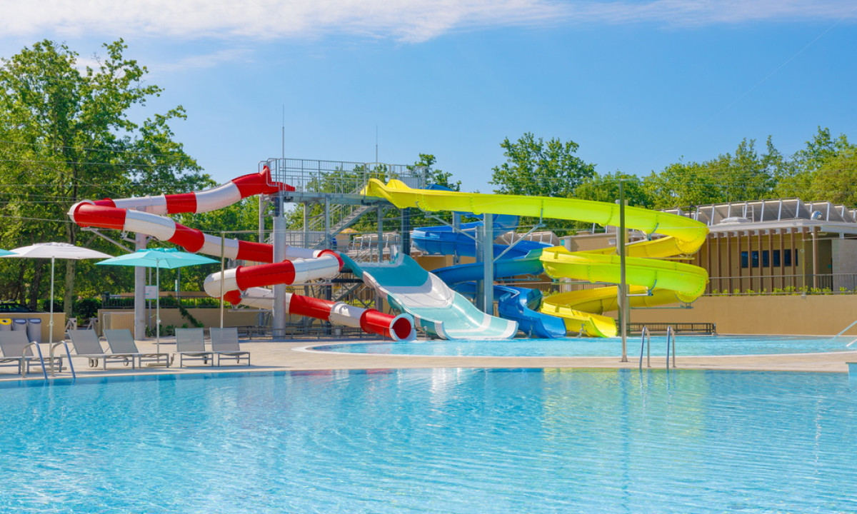 Waterslides curl above a large, clear swimming pool with lounge chairs and umbrellas beside wooded surroundings at Bijela Uvala