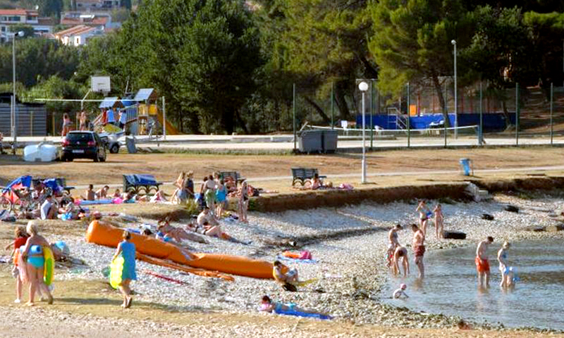 People are swimming, sunbathing, and playing on a rocky lakeshore with trees, benches, and a children's playground nearby at Stupice