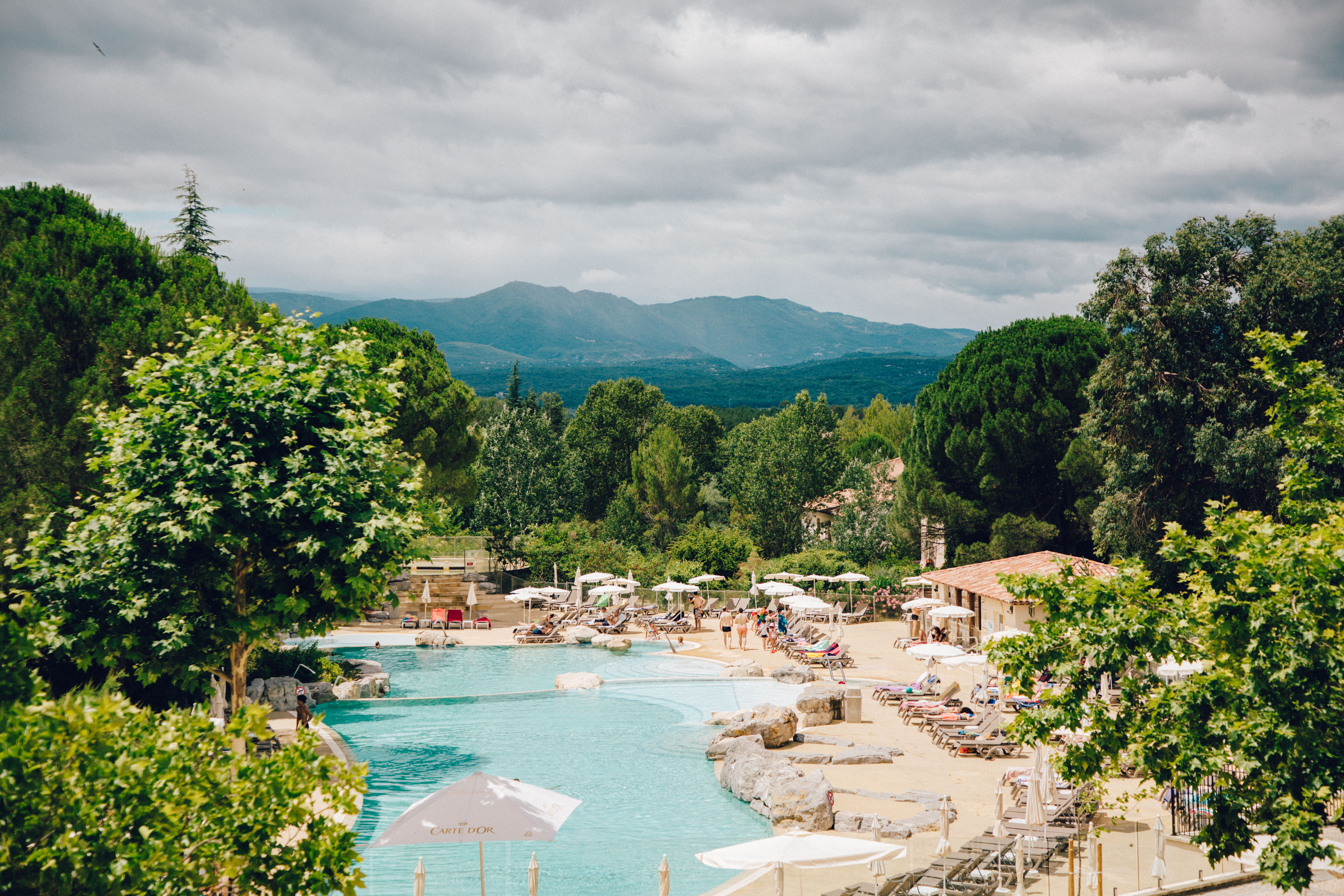 Swimming pool surrounded by lounge chairs and trees, with mountains in the background at Le Rouret en Ardeche