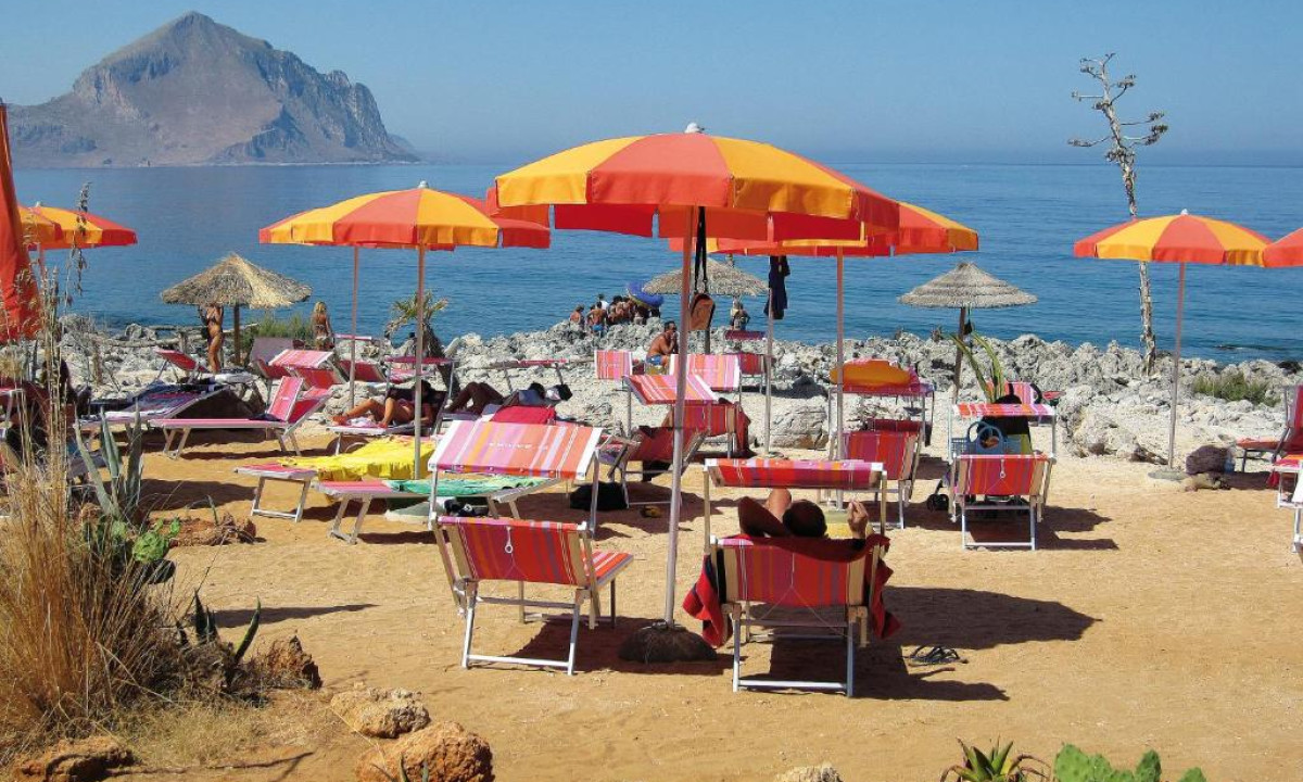 Colorful beach umbrellas shade lounge chairs on a sandy shore by a rocky coastline, with a mountain in the background at El Bahira