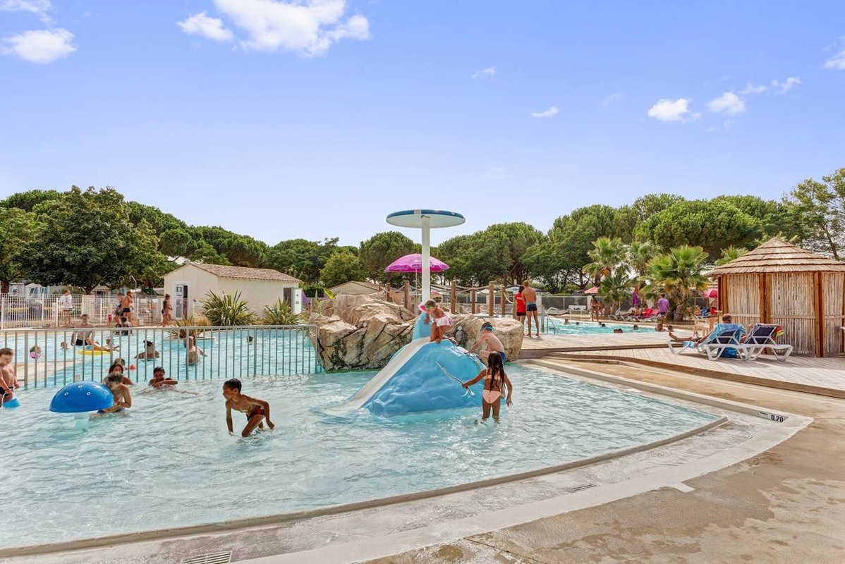 Children playing in a shallow pool with slides and water features, surrounded by trees and lounge chairs at Bon Port