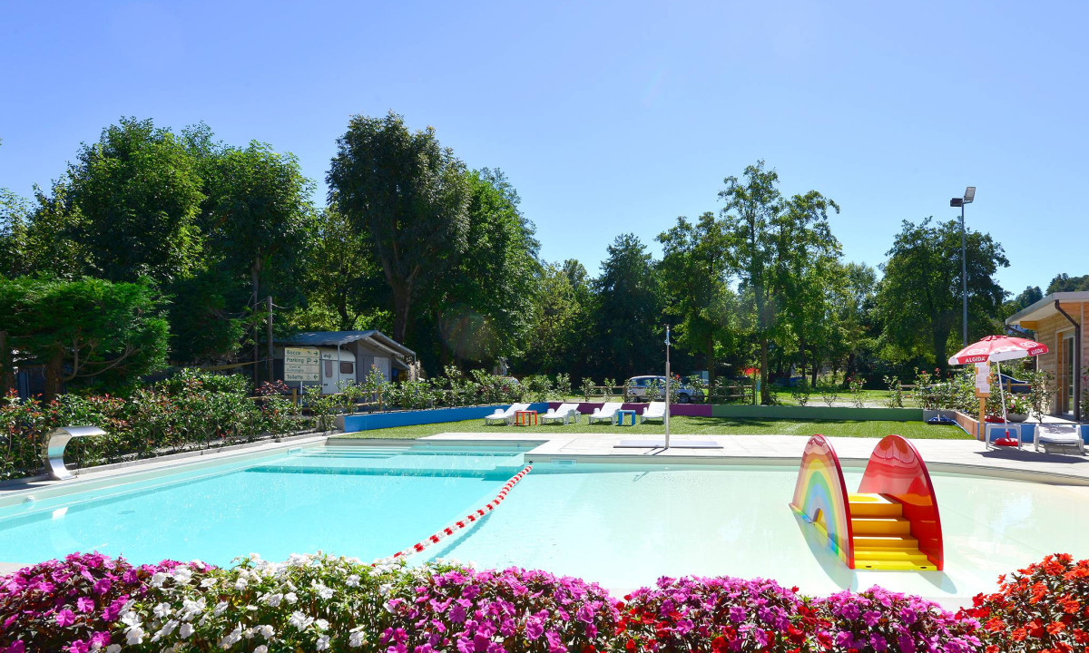 Swimming pool with rainbow slide, surrounded by colorful flowers and lounge chairs, set in a lush, green park at Eden Maggiore