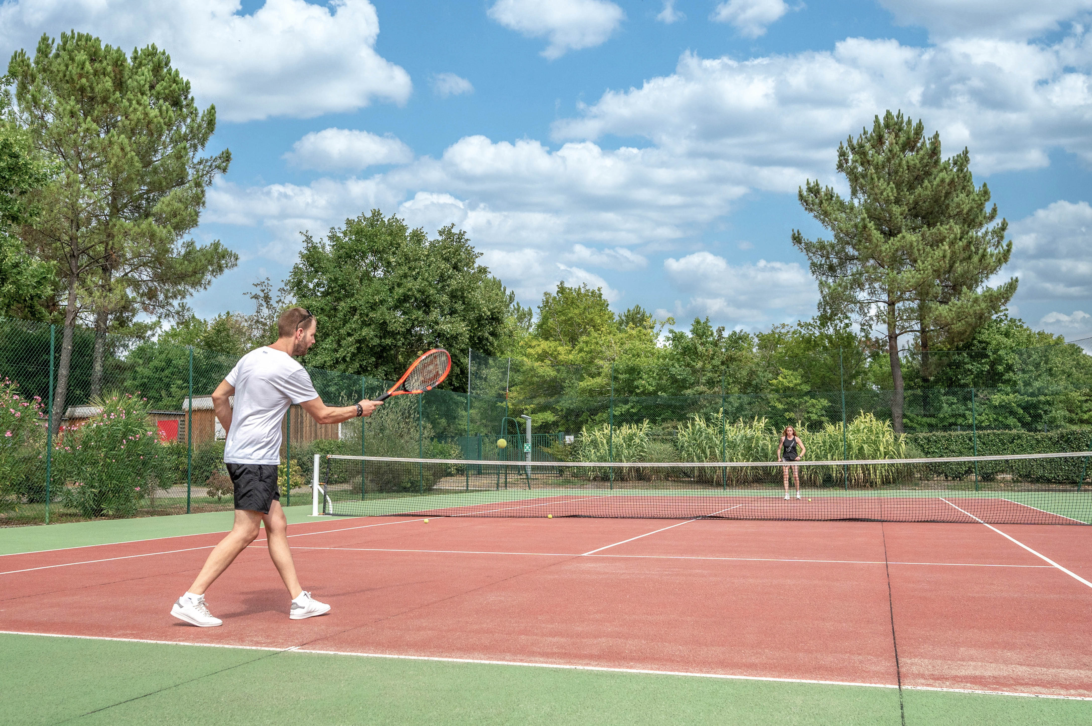 Person playing tennis, hitting ball on an outdoor court surrounded by trees and greenery at Au Lac de Biscarrosse