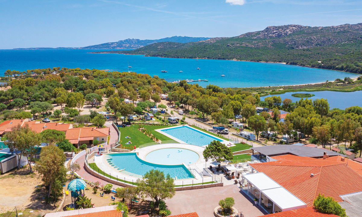 The pool is surrounded by red-roofed buildings and green trees, overlooking a campsite near a blue bay at Capo D'Orso