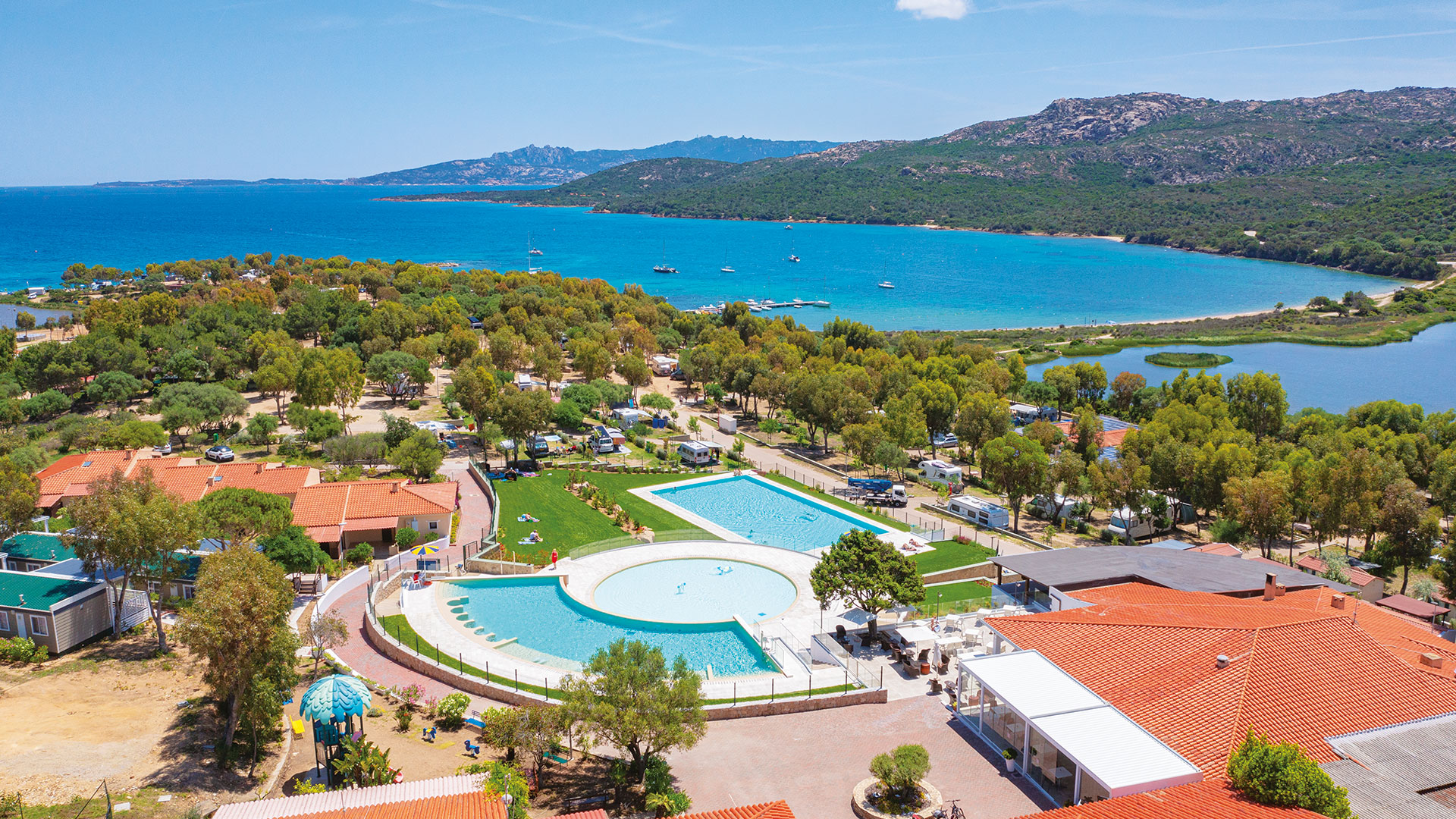 The pool is surrounded by red-roofed buildings and green trees, overlooking a campsite near a blue bay at Capo D'Orso
