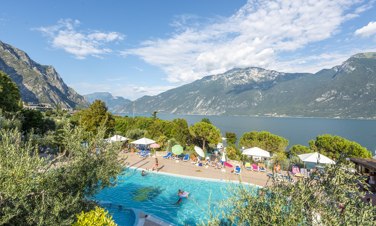 Swimming pool with people relaxing and playing, surrounded by sunbeds and umbrellas, overlooking a mountainous lake view at Garda