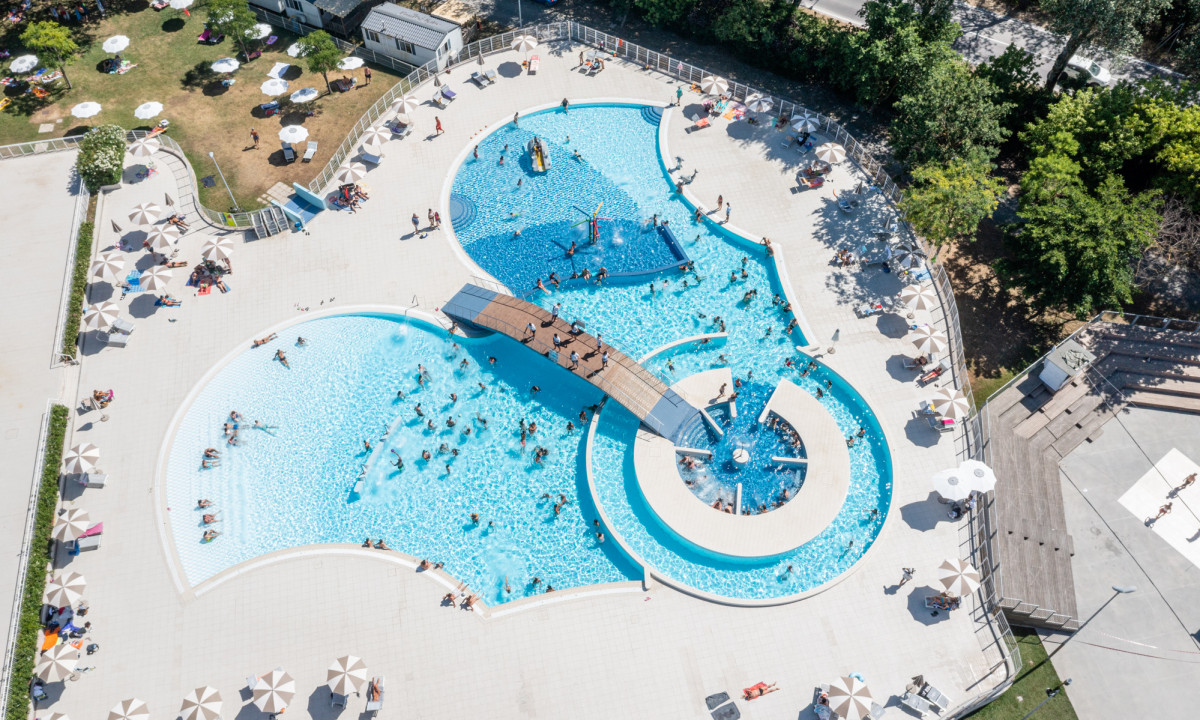 People swimming in interconnected pools with a bridge, surrounded by loungers and umbrellas at Adriano Family Camping Village