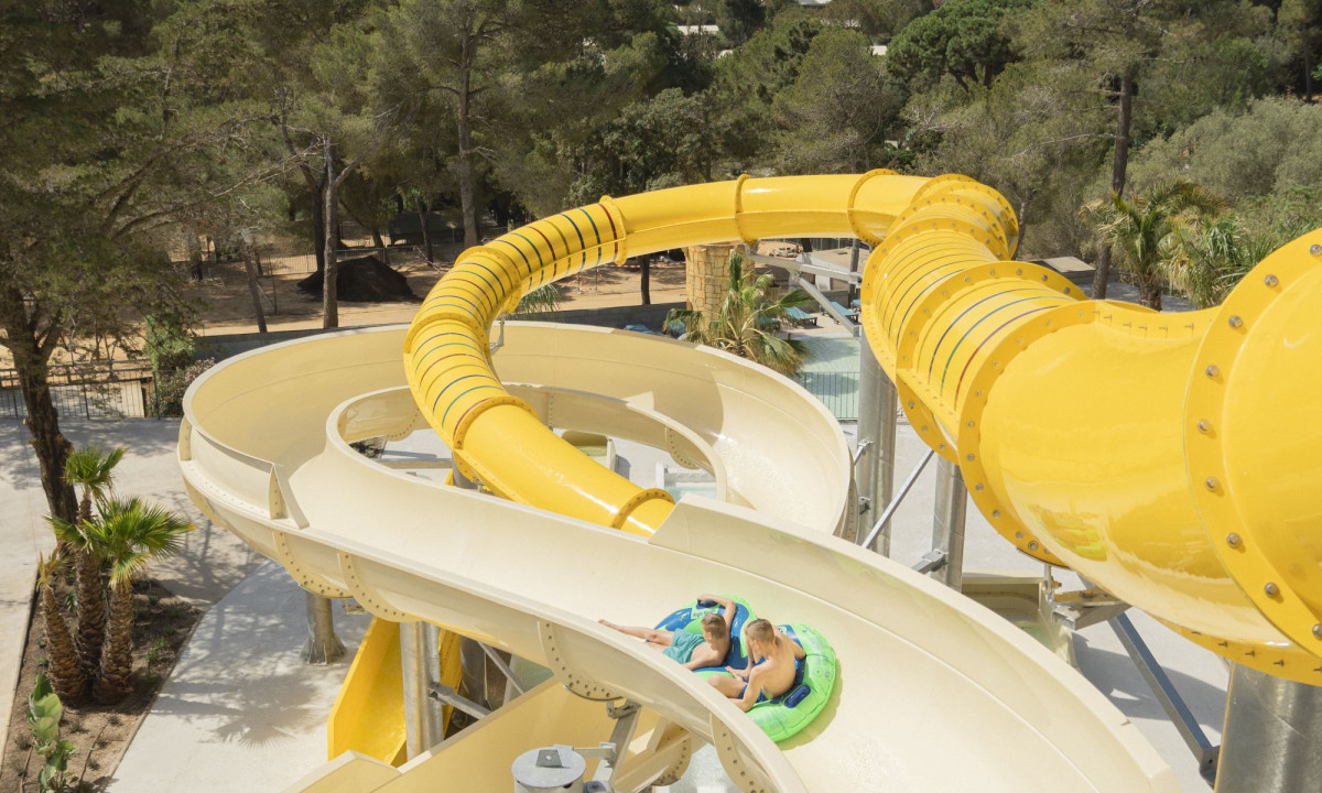 Two people riding a curving water slide on an inflatable tube, surrounded by lush trees at Internacional de Calonge