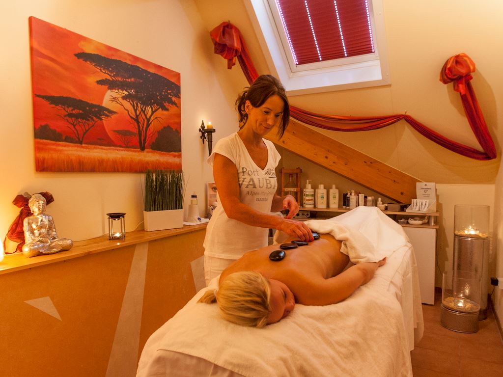 A woman administers a hot stone massage to a client on a massage table, with a cozy spa room setting at Landal Dwergter Sand