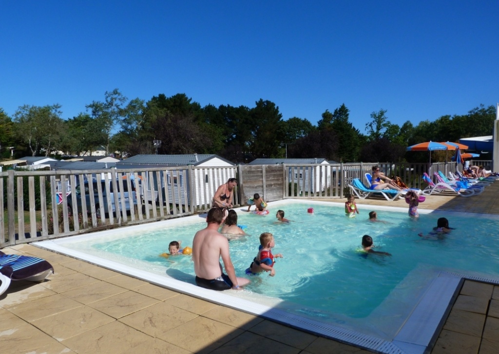 Children and adults are swimming in a pool with several people lounging on pool chairs under umbrellas, surrounded by nature and cabins at Domaine de Bréhadour