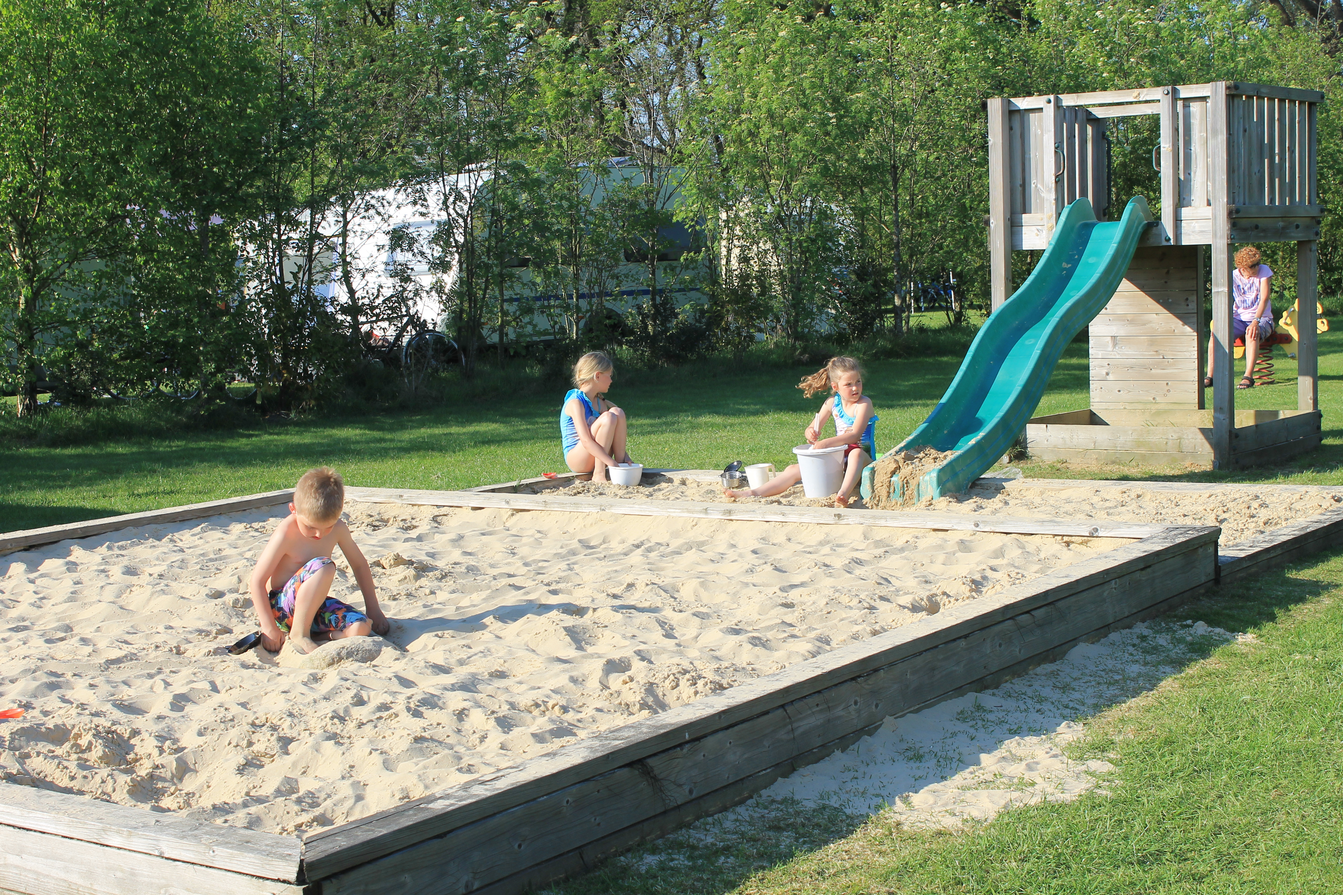 Children playing in a sandbox with a slide beside them, surrounded by greenery and trees at Dal van de Mosbeek