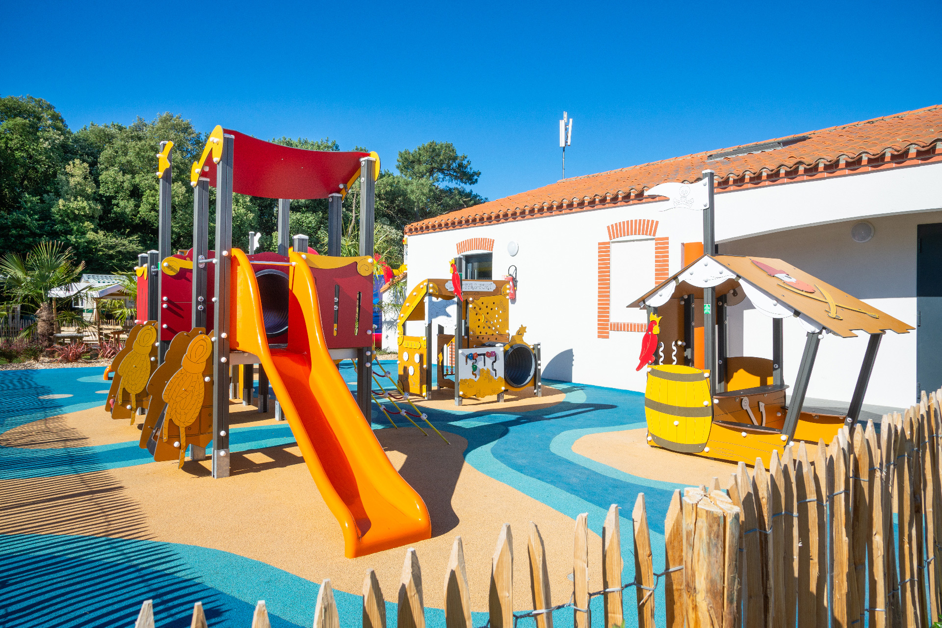 Brightly colored playground set with slides and climbing structures, surrounded by greenery and adjacent to a white building at Le Littoral