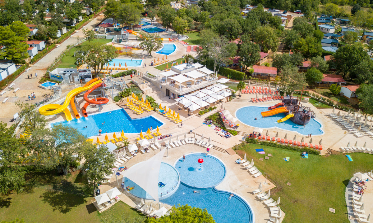 Aerial view of multiple pools with slides and sun loungers surrounded by greenery, cabins, and trees at Lanterna