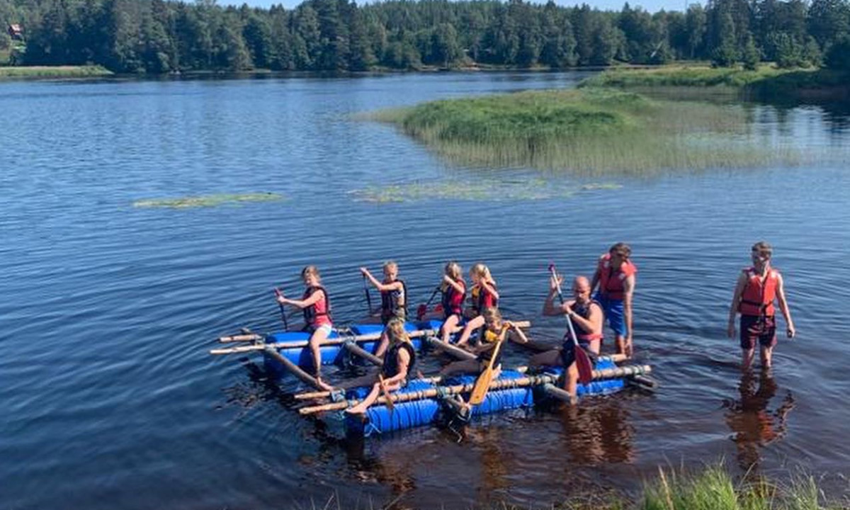 Children wearing life vests paddle a homemade raft on a lakeside, amidst a forested environment at Yttermalungs