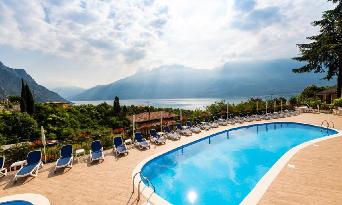 Blue pool surrounded by lounge chairs, overlooking a scenic lake and mountain landscape under a partly cloudy sky at Residence Oasi