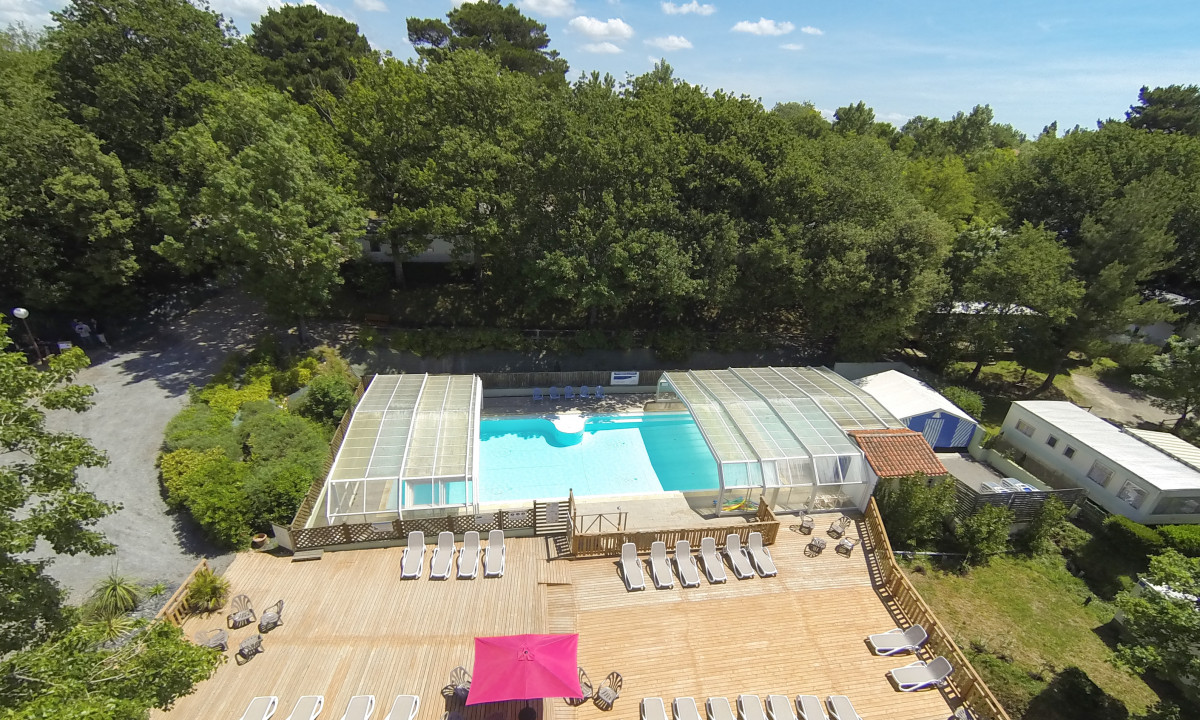 Swimming pool with a retractable cover beside a wooden deck with lounge chairs in a forested campground area at Le Paradis