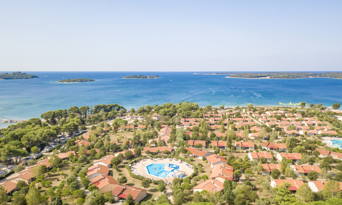 Aerial view of a coastal campsite with bungalows arranged around a pool, surrounded by trees and sea at Bi-Village