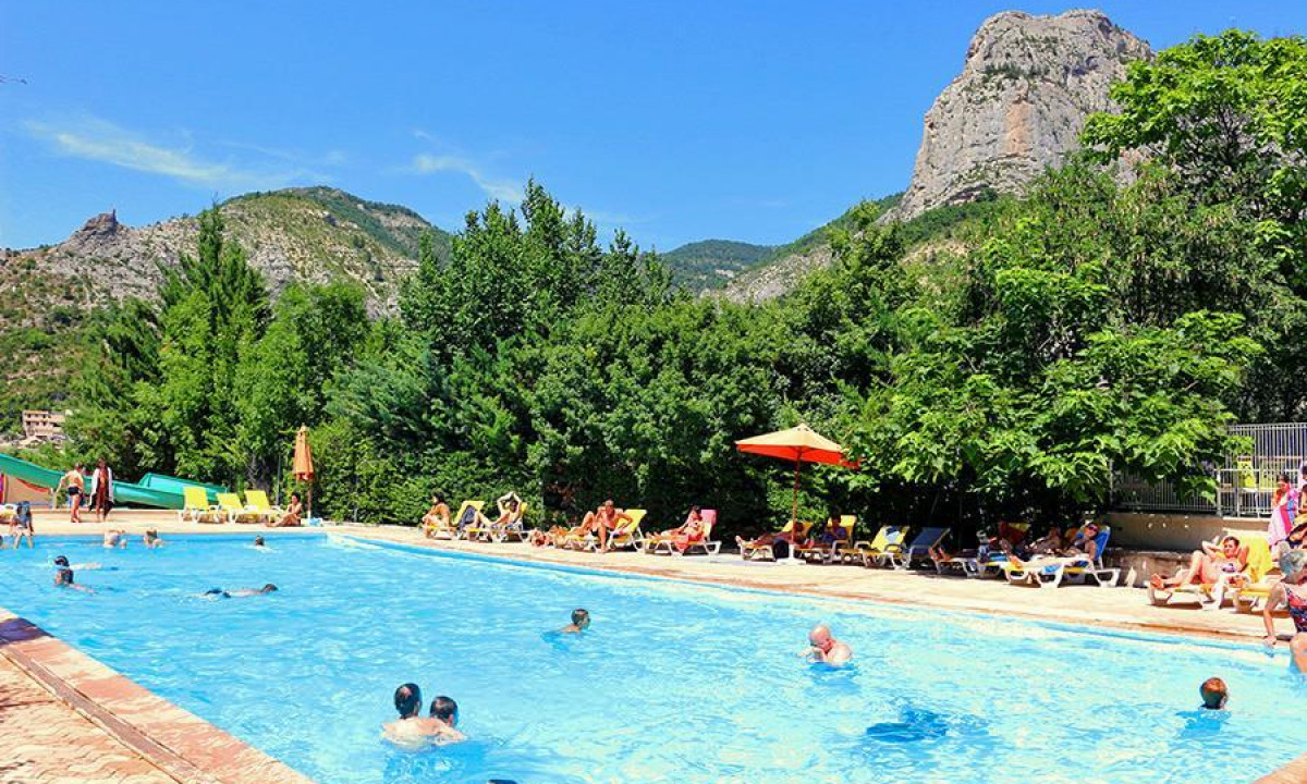 People swimming in a crowded pool surrounded by lounge chairs, trees, and mountains in the background at Princes d'Orange