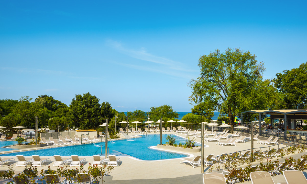 Outdoor swimming pool surrounded by lounge chairs and umbrellas, with trees and a restaurant in the background at Aminess Maravea Camping Resort