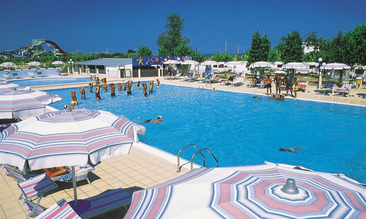 People swim and exercise in a large outdoor pool surrounded by umbrellas, sunbeds, and greenery at Stella del Sud