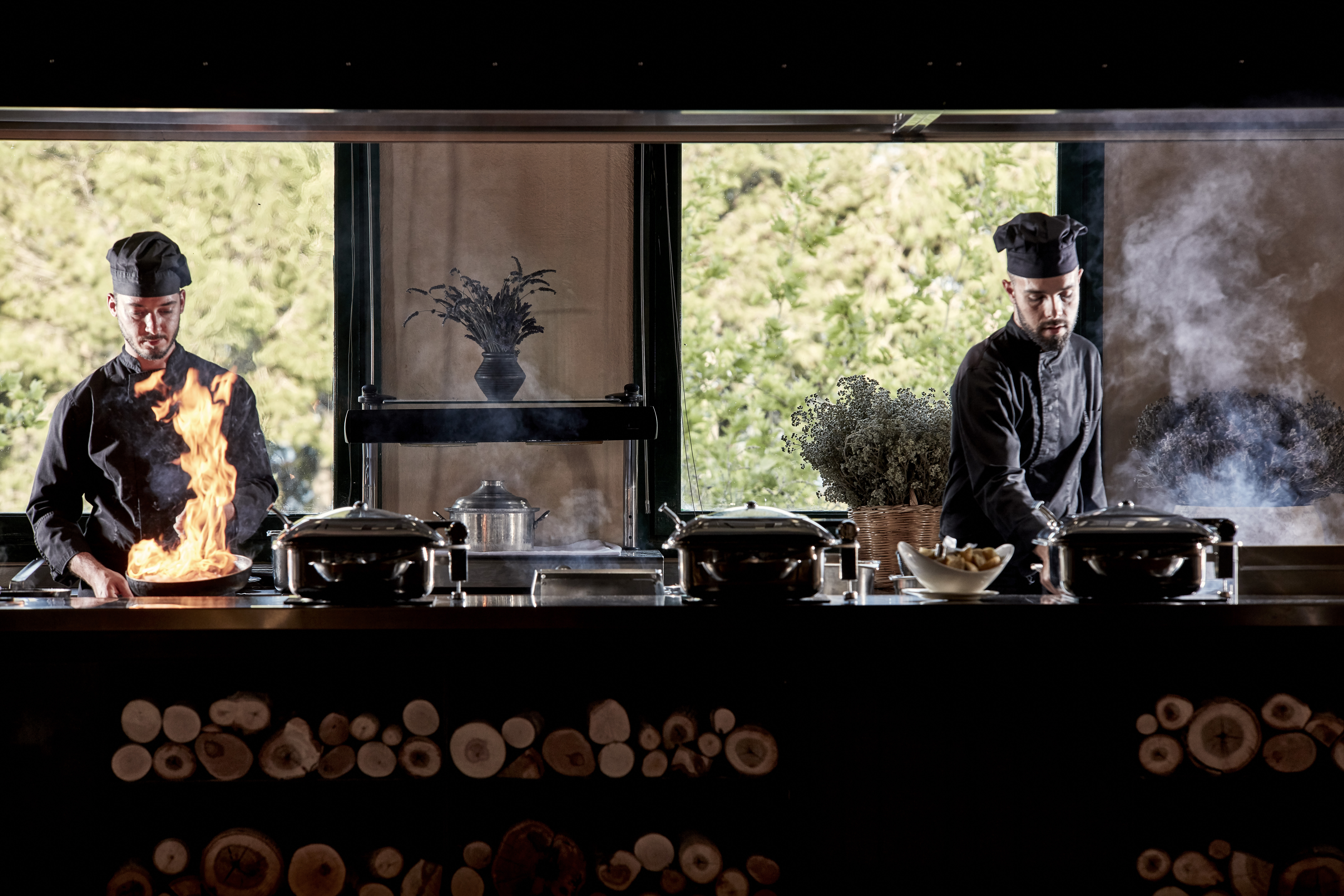 Chefs cooking with flames and smoke in an open kitchen with wooden accents, trees visible through large windows at Candia Park Village