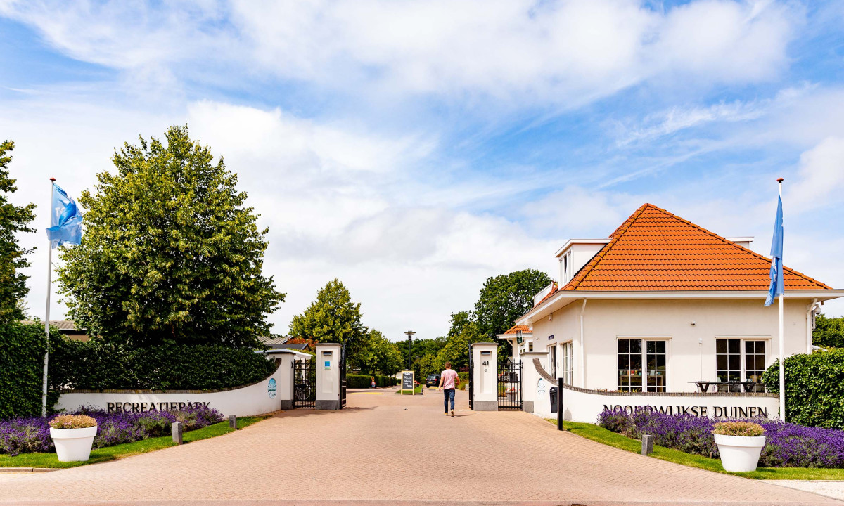 Entrance gate leading to a recreational park, trees and buildings with orange roofs in the background at Europarcs Noordwijkse Duinen