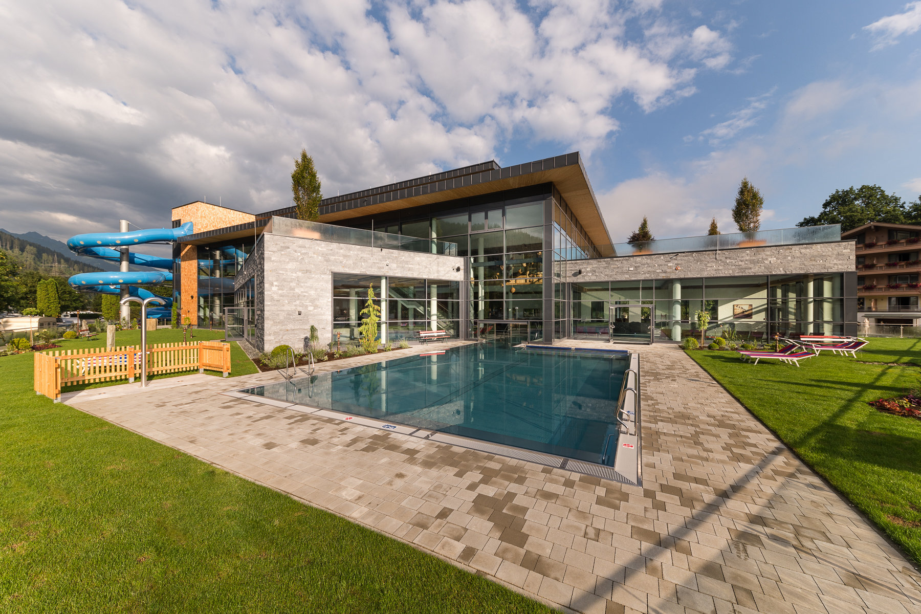 Swimming pool surrounded by modern building, water slide, and lounge chairs on a sunny day at Sportcamp Woferlgut