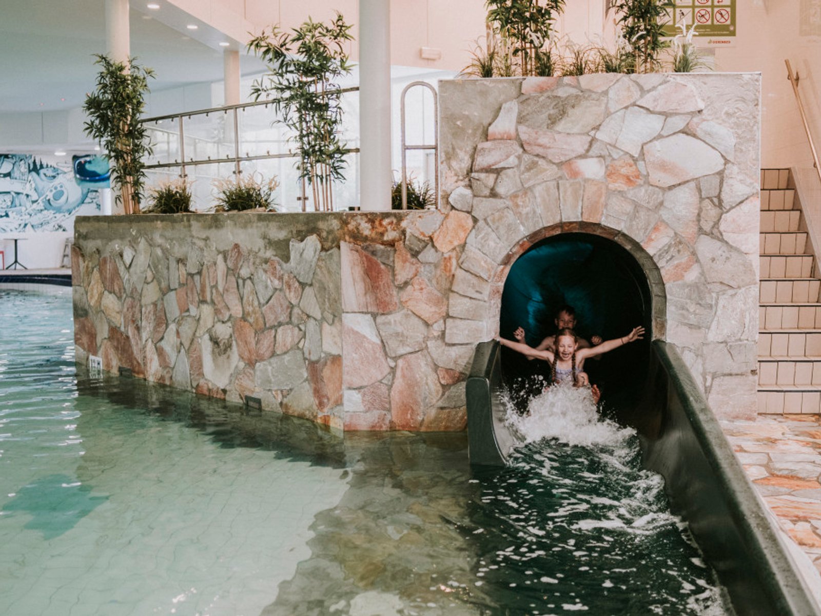 Children slide down a dark water slide, splashing into a pool surrounded by stone walls, indoors at Vakantiepark Dierenbos
