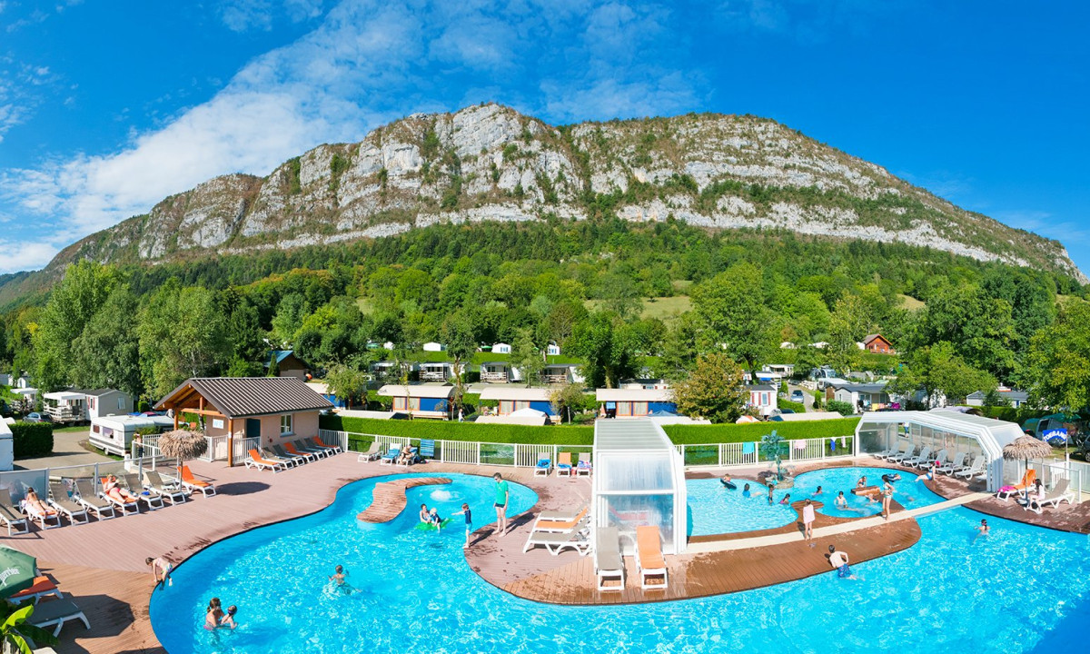 Swimming pool with people relaxing and swimming, surrounded by trees, cabins, and mountains in the background at Les Fontaines