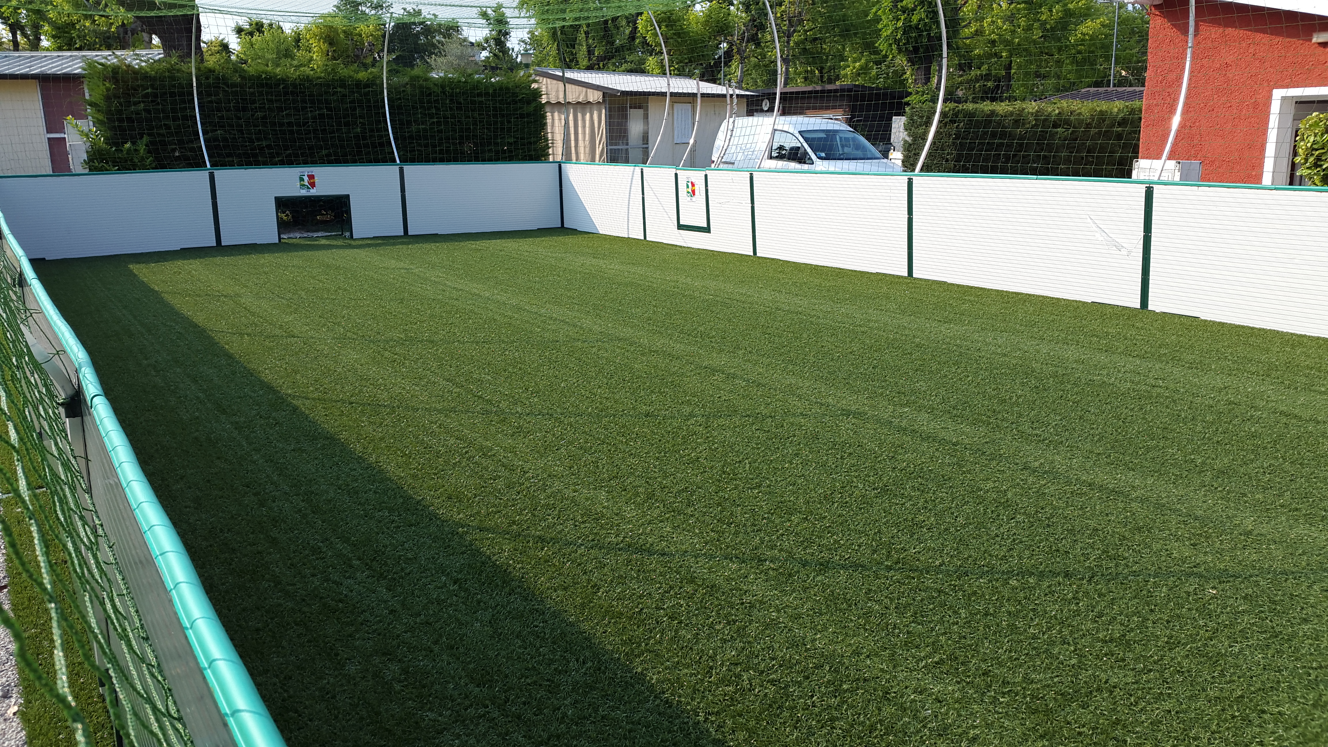 Artificial turf soccer field with netted fencing, surrounded by trees and buildings at Camping Sereno