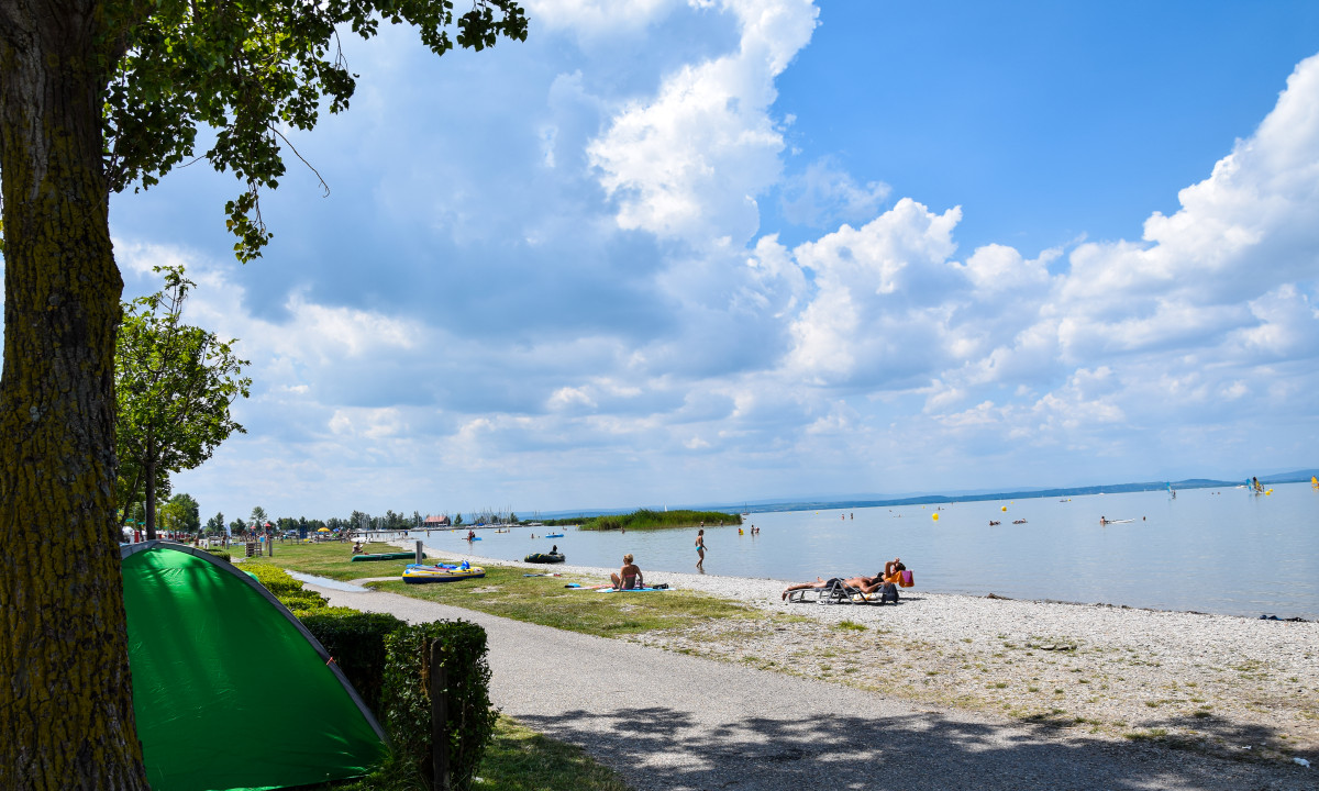 A green tent stands under trees near a lakeside with people relaxing and swimming on a sunny day at Podersdorf