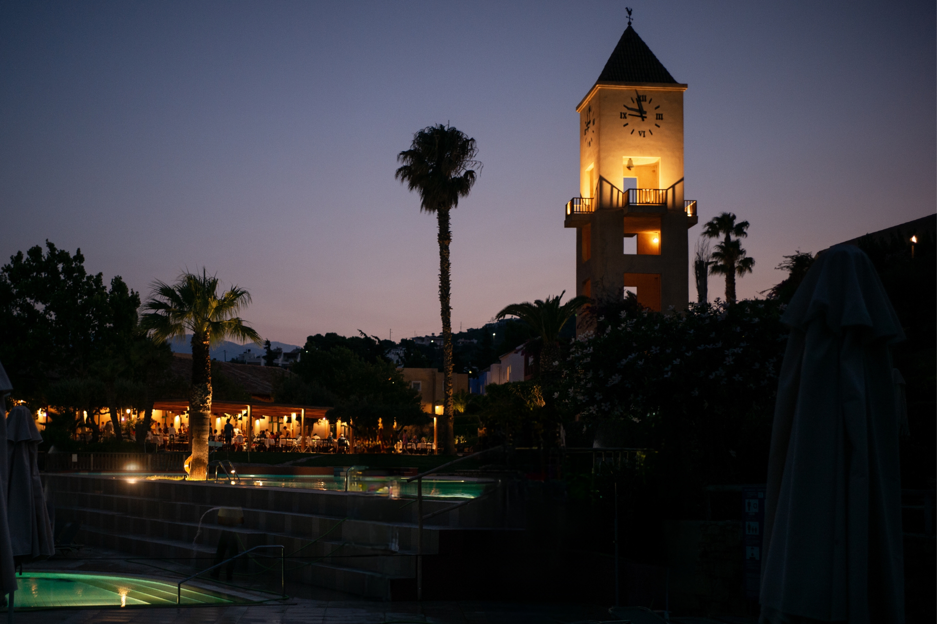 Illuminated clock tower standing amidst palm trees and buildings at dusk, overlooking a pool area at Candia Park Village