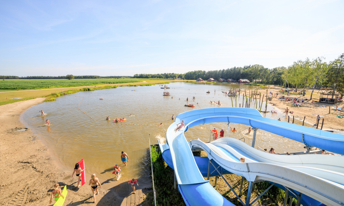 Waterslide flowing into a busy lakeside swimming area with people playing on sandy beach and grassy fields at de Flaasbloem
