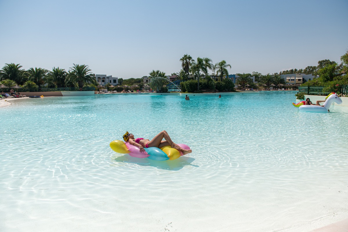 Person relaxing on an inflatable lounger in a large, clear blue pool surrounded by palm trees and resort buildings at I Turchesi Club Village