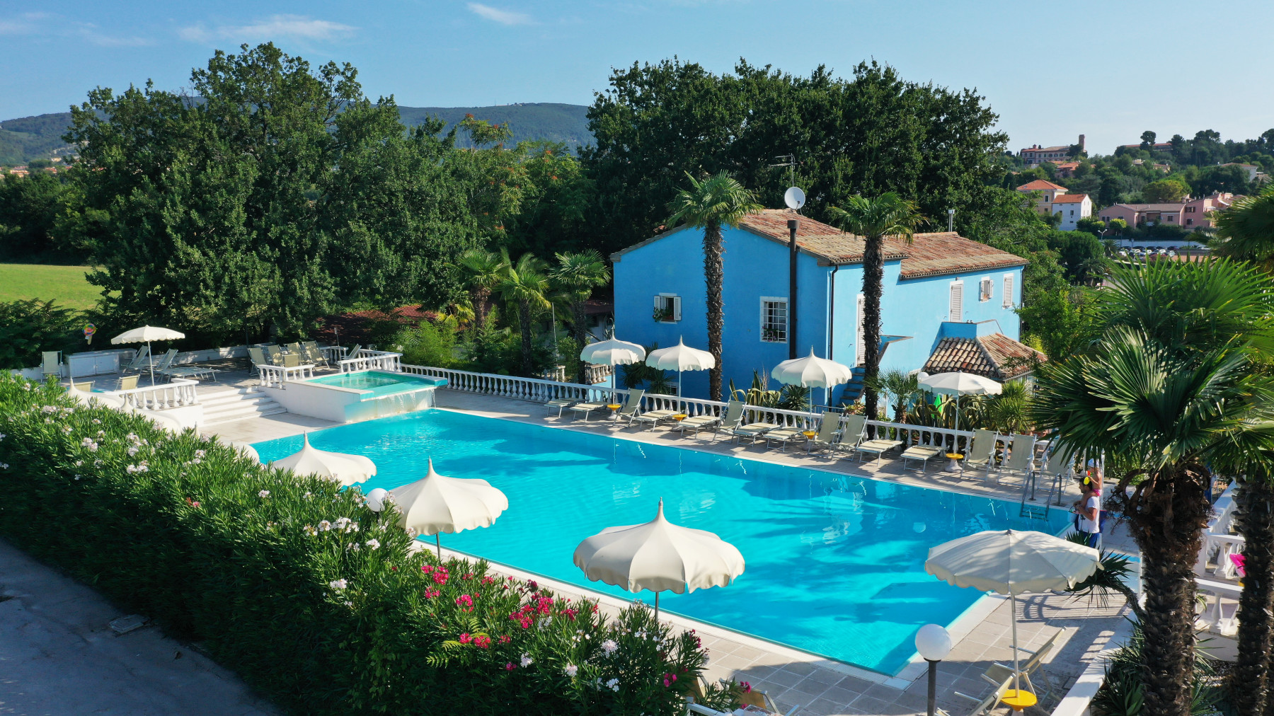 A blue swimming pool surrounded by umbrellas and lounge chairs, next to a vibrant blue house amidst greenery at Riviera Village